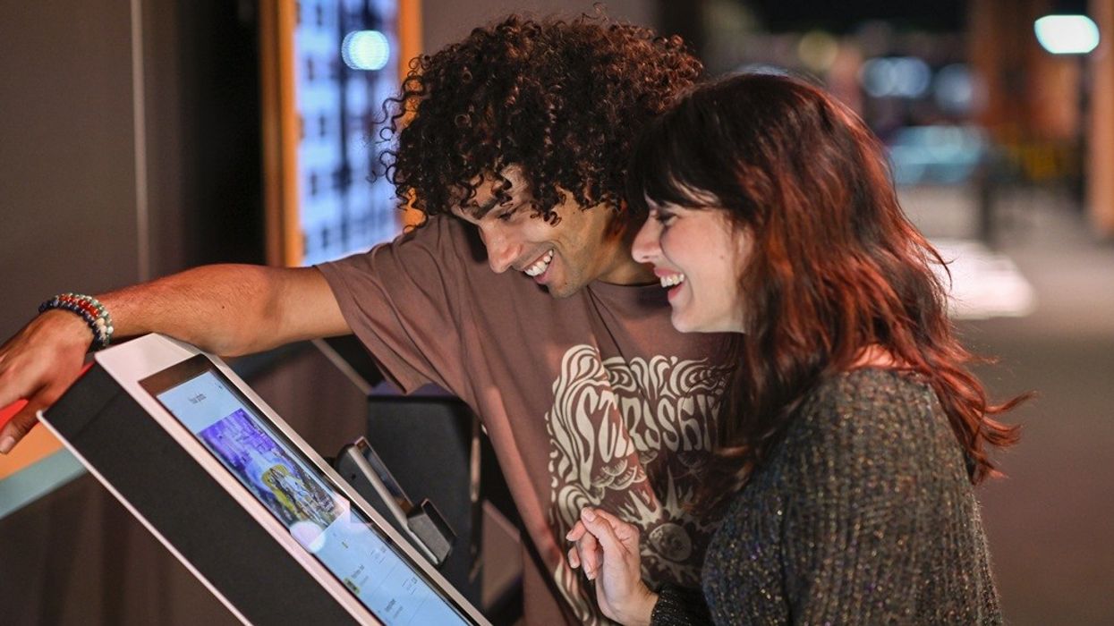 A couple smiles while using a touchscreen kiosk indoors.