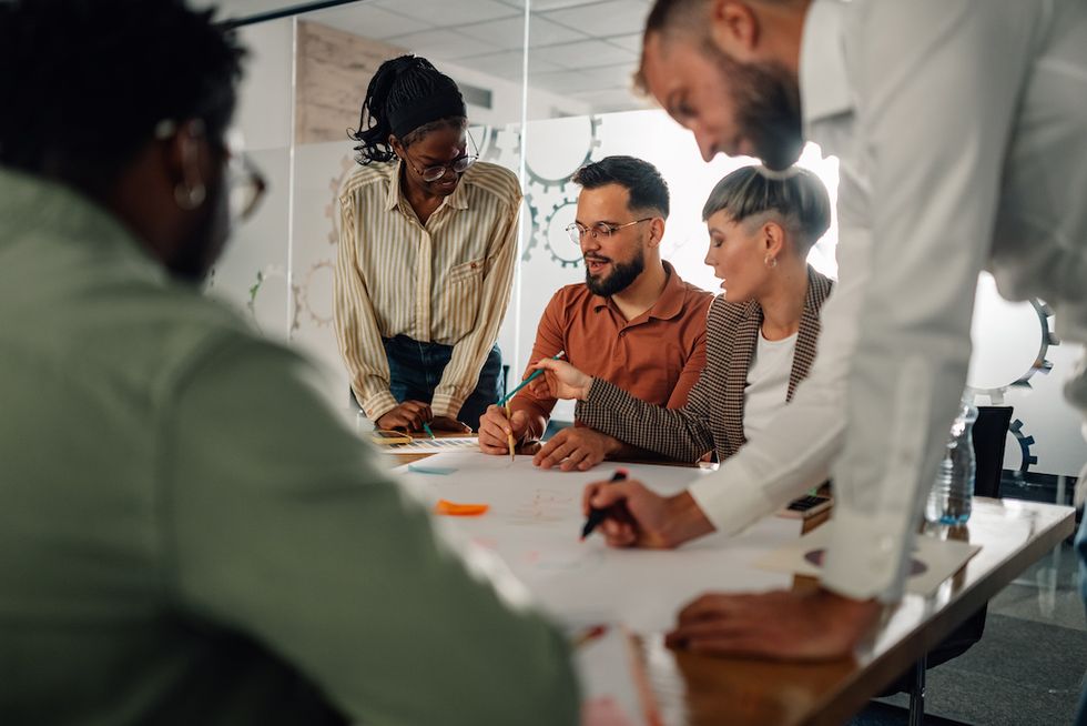 A diverse group collaborates at a table, working on a large sheet of paper.