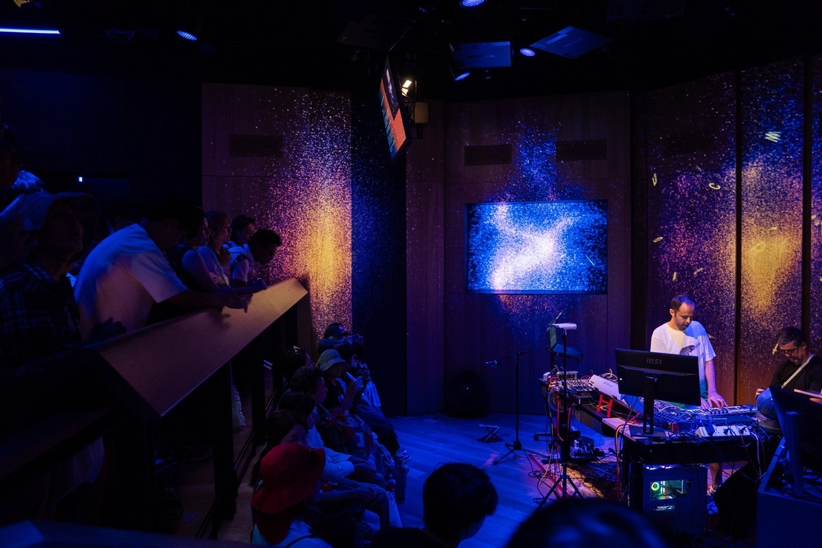 A DJ performs for a seated audience in a dimly lit room with vibrant, cosmic visuals.