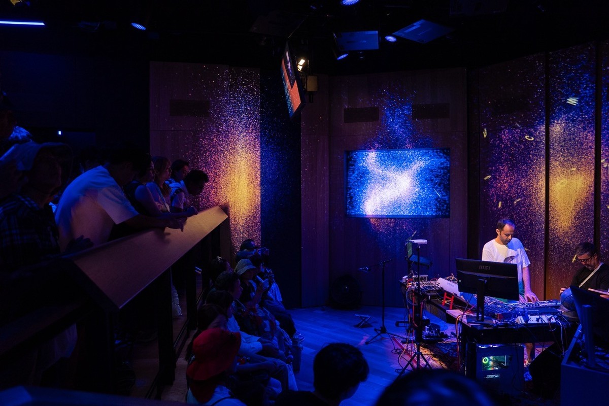 A DJ performs for a seated audience in a dimly lit room with vibrant, cosmic visuals.