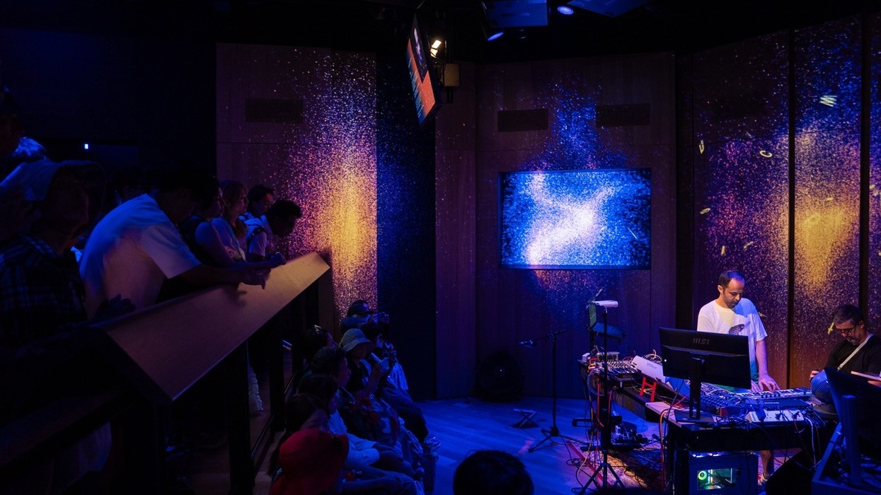 A DJ performs for a seated audience in a dimly lit room with vibrant, cosmic visuals.