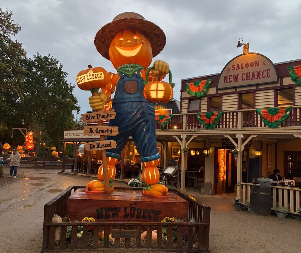 A giant smiling pumpkin cowboy holds a lantern outside a Western-themed saloon.