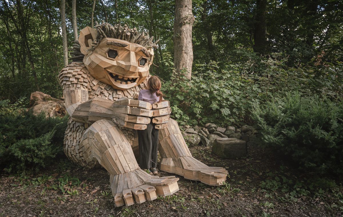 A girl hugs a large wooden troll sculpture in a forest setting.