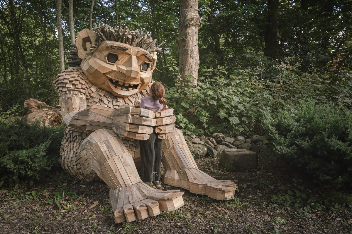 A girl hugs a large wooden troll sculpture in a forest setting.
