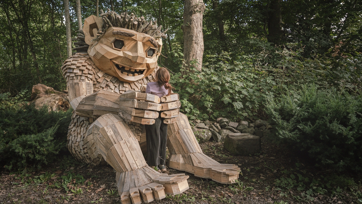 A girl hugs a large wooden troll sculpture in a forest setting.
