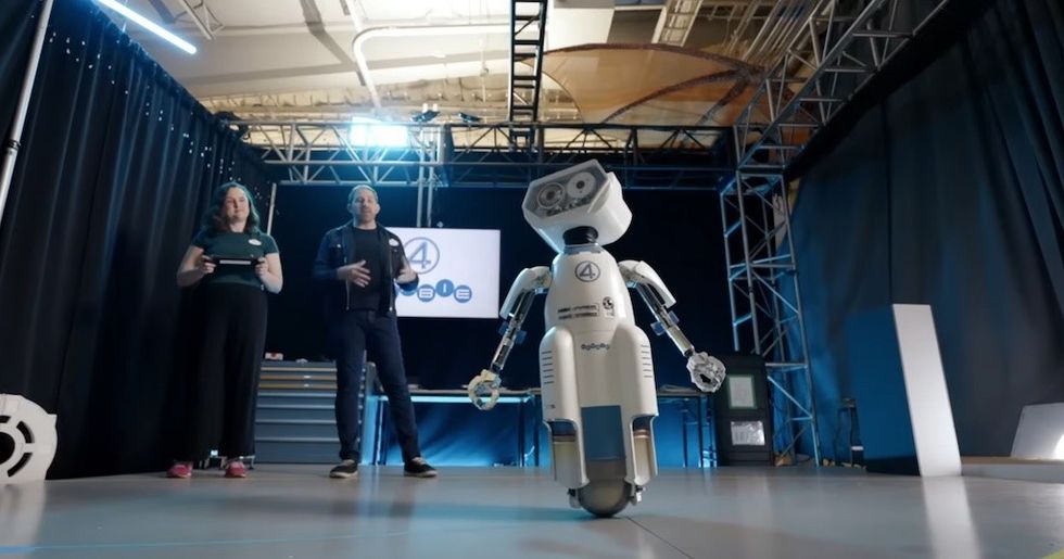 A humanoid robot on a stage with two people and a screen in the background.