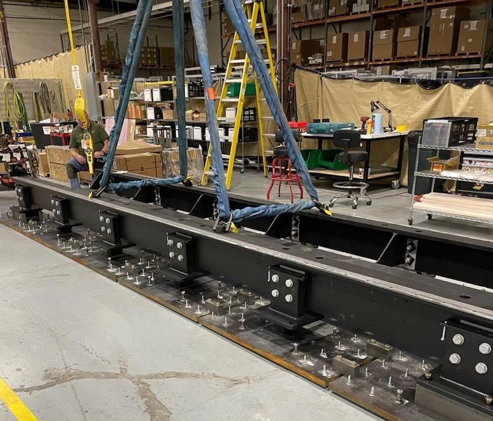 A large metal beam suspended by straps in an industrial workshop setting.