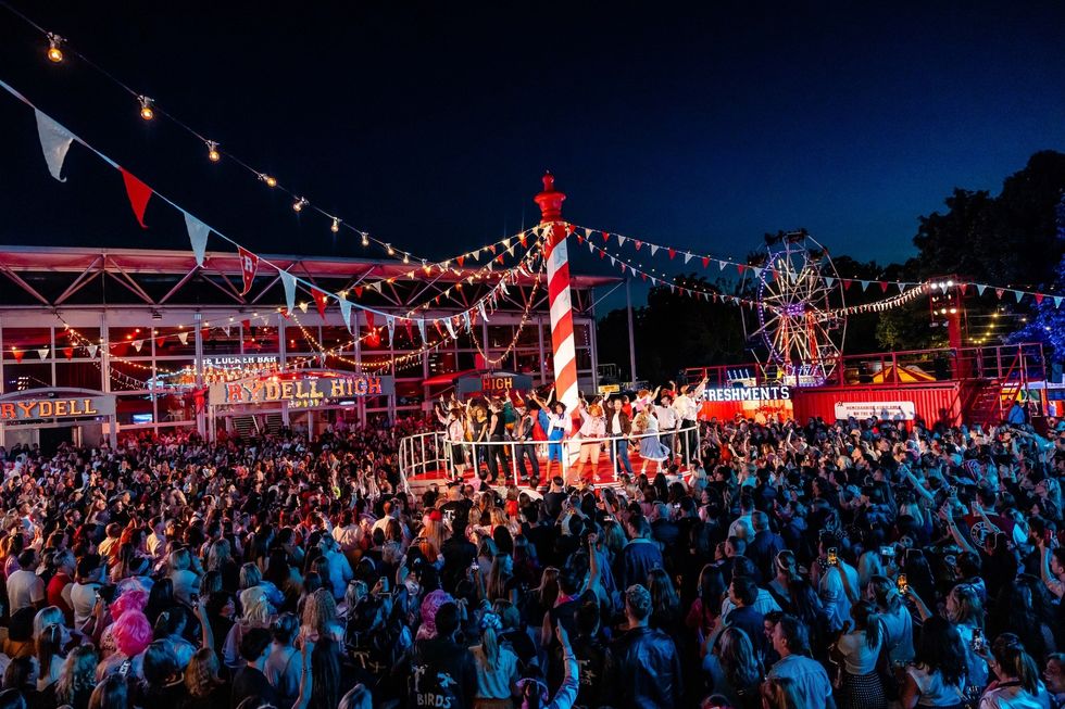 A lively outdoor party with a large crowd, stage performance, and festive lights at night.