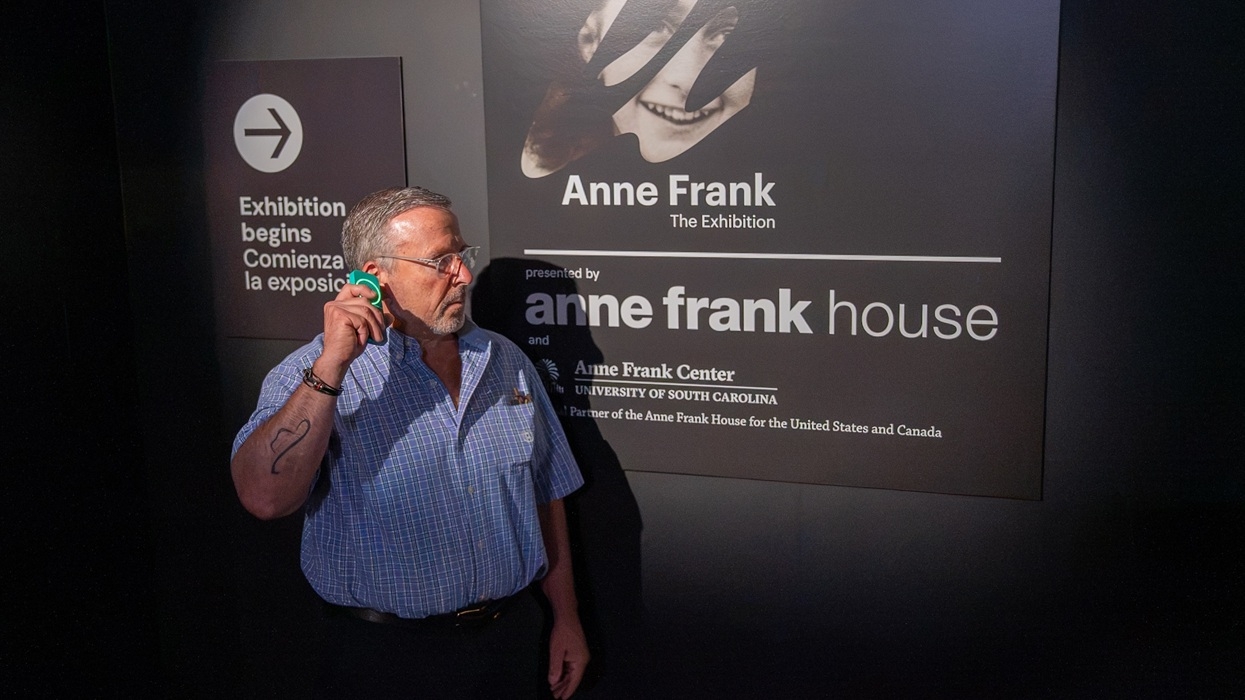 A man stands beside an Anne Frank exhibition sign, holding up a Guide-ID audio tour device.