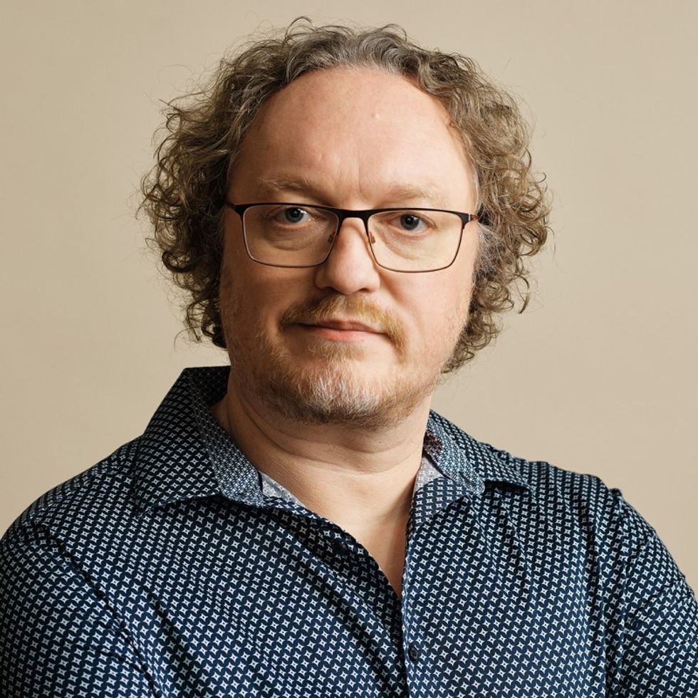 A man with curly hair, glasses, and a patterned shirt against a plain background.