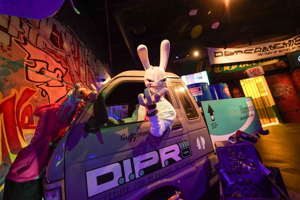 A person in a rabbit mask poses from a graffitied van in a dimly lit, colorful room at Dopeameme