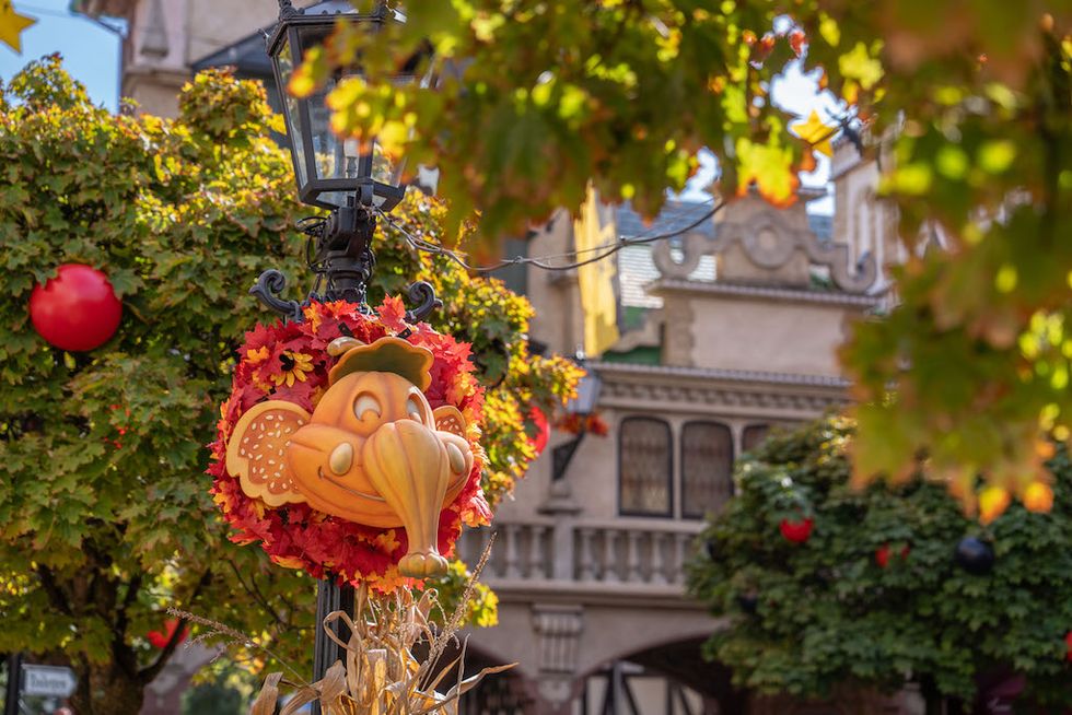 A pumpkin-faced autumn wreath hangs from a lamppost amid colorful fall foliage.