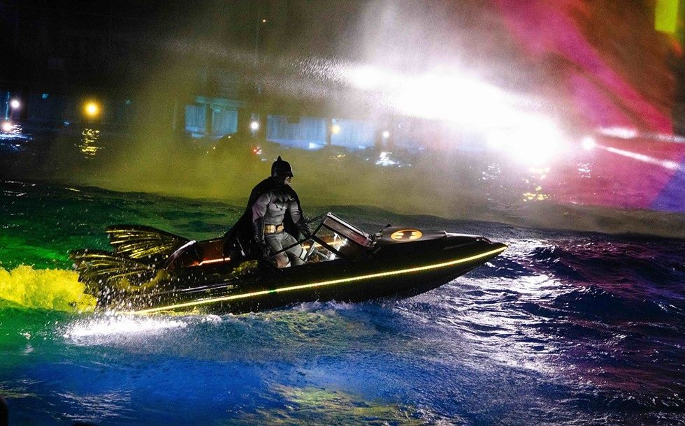 A superhero speeds on a themed boat in colorful, illuminated water at night.