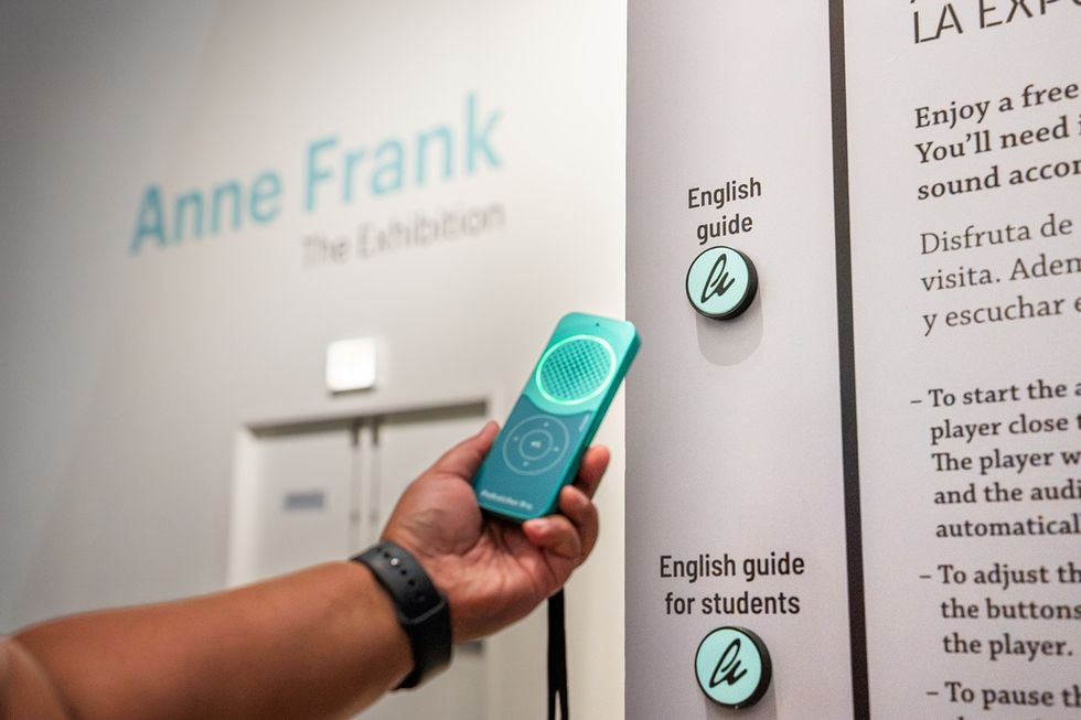 A visitor holds a turquoise Guide-ID audio guide beside English guide buttons at the Anne Frank exhibition.