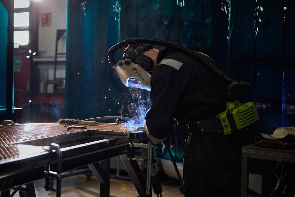 A welder in protective gear welding metal at a workbench, with sparks flying.