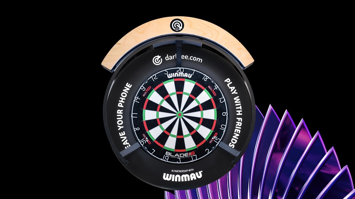 A Winmau dartboard mounted in a black surround with a Dartsee Mini unit with light wood top, against a black and purple background.