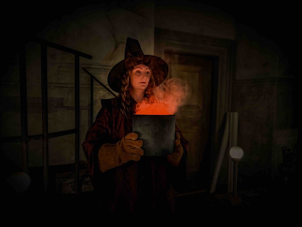 A witch in a dark room holds a steaming, glowing cauldron.