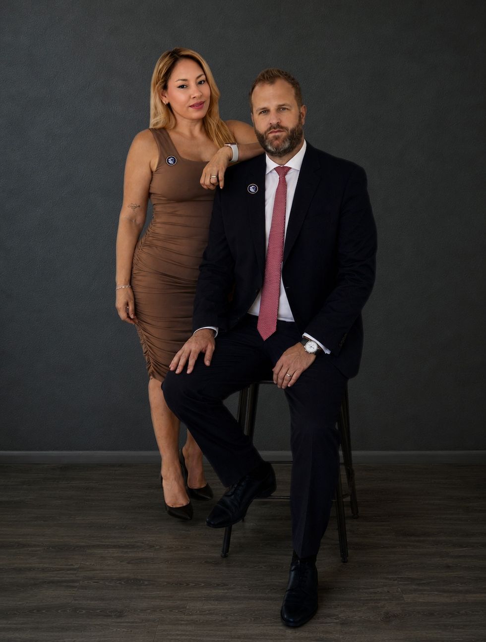 A woman and man in formal attire pose confidently against a dark background.