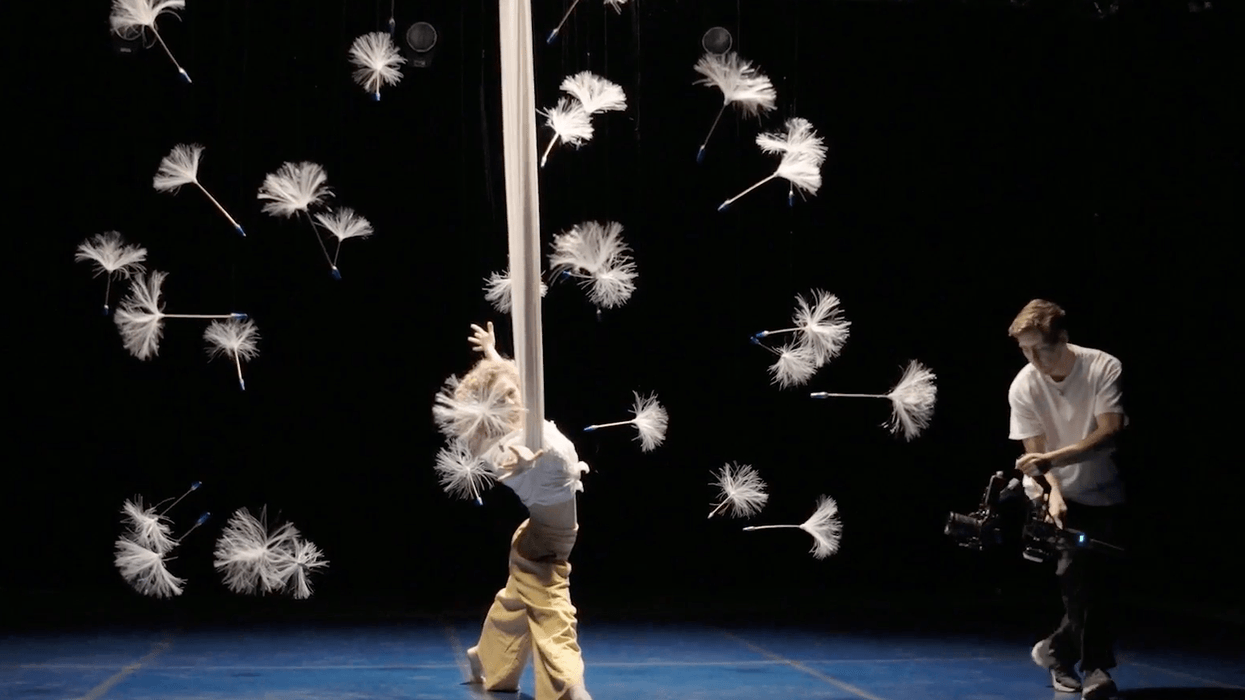 Aerial performer spins amid floating dandelions; cameraman films on a dark stage.