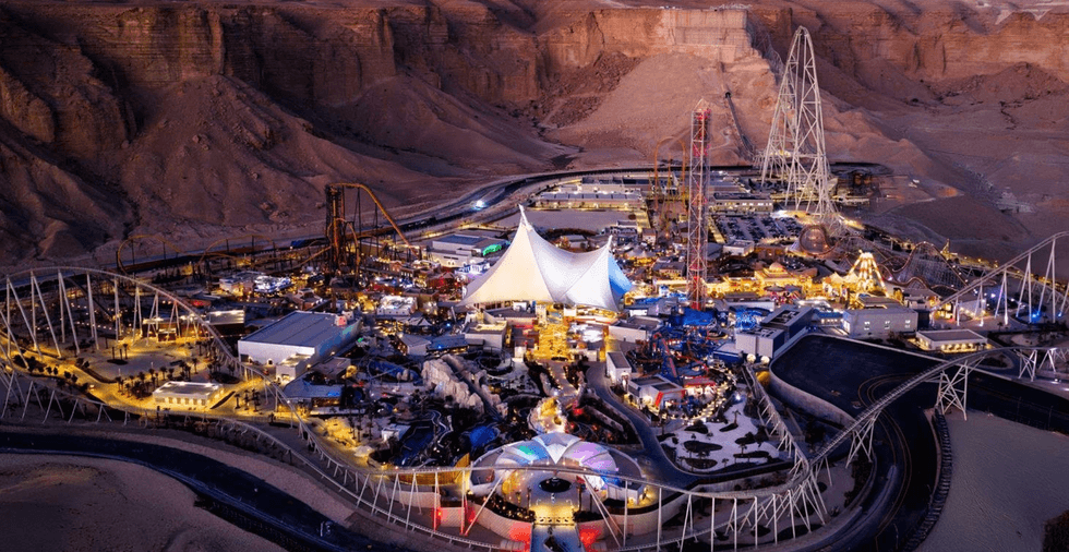 Aerial view of a brightly lit desert amusement park with roller coasters at dusk.