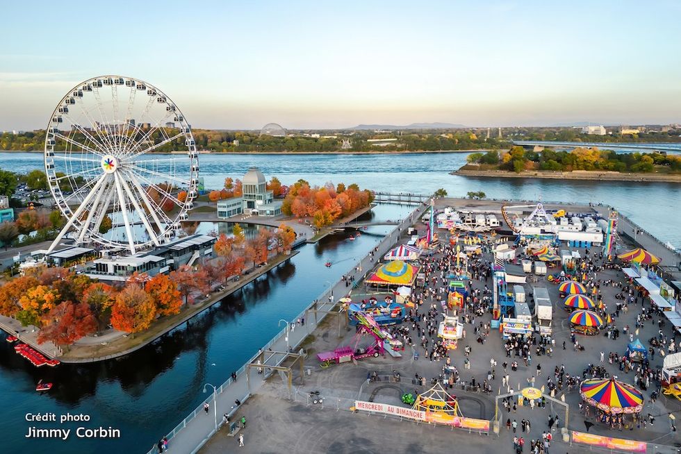Aerial view of a colorful riverside fair with Ferris wheel and autumn trees.