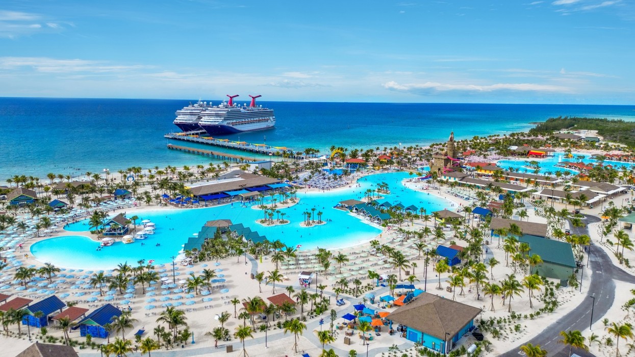 Aerial view of a cruise ship docked near a beach resort with pools and palm trees.