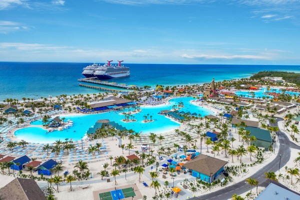 Aerial view of a cruise ship docked near a beach resort with pools and palm trees.