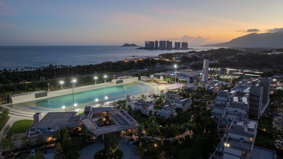 Aerial view of a lit-up surf pool and buildings by the sea at sunset at Riyue Bay Surf Resort