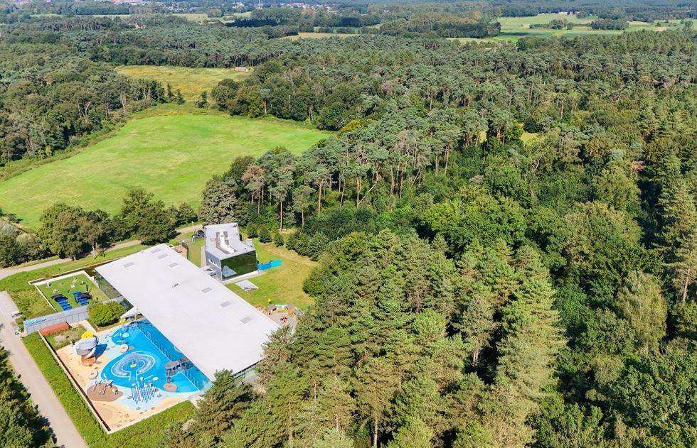 Aerial view of a swimming pool complex amid lush, dense forest and open green fields.