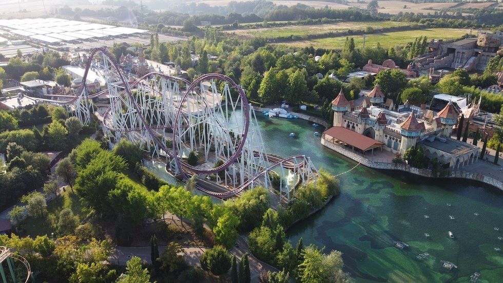 Aerial view of a theme park with a large roller coaster and a castle by a green lake.