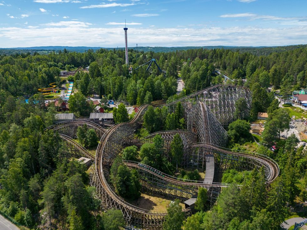 Aerial view of a wooden roller coaster surrounded by lush green trees in a theme park.