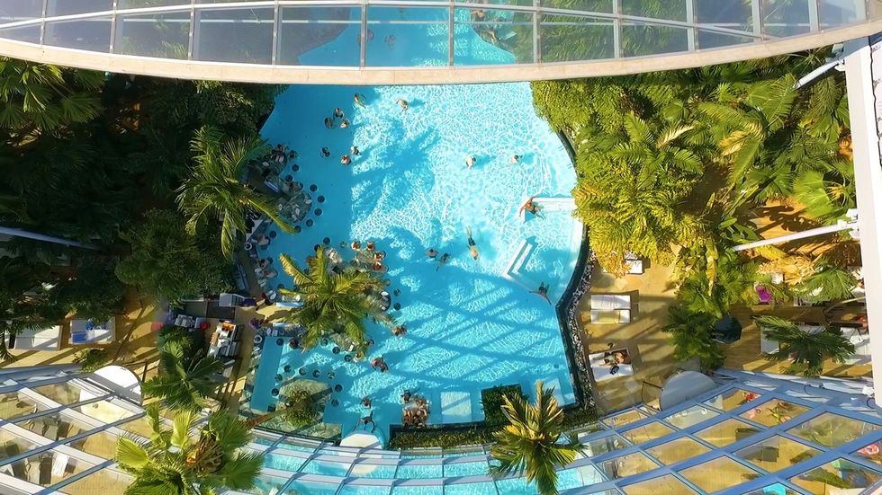 Aerial view of indoor tropical pool surrounded by lush greenery and people swimming.