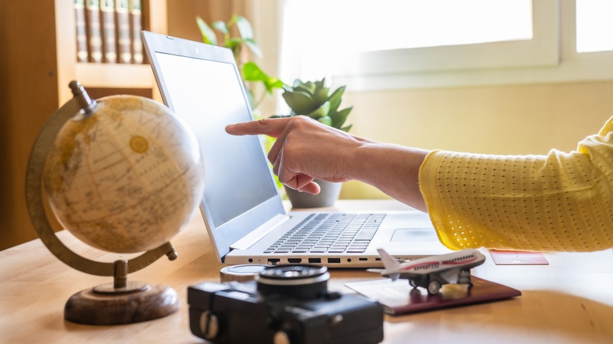 AI search Woman pointing at laptop screen while planning a trip on a booking website, surrounded by a globe, camera, passport, and airplane model on the desk, creating an organized itinerary