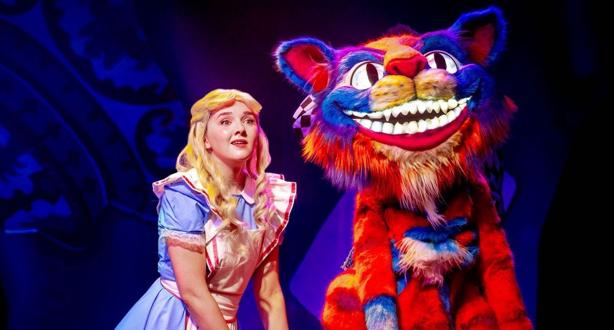 Alice in Wonderland beside a colorful, grinning Cheshire Cat on stage.
