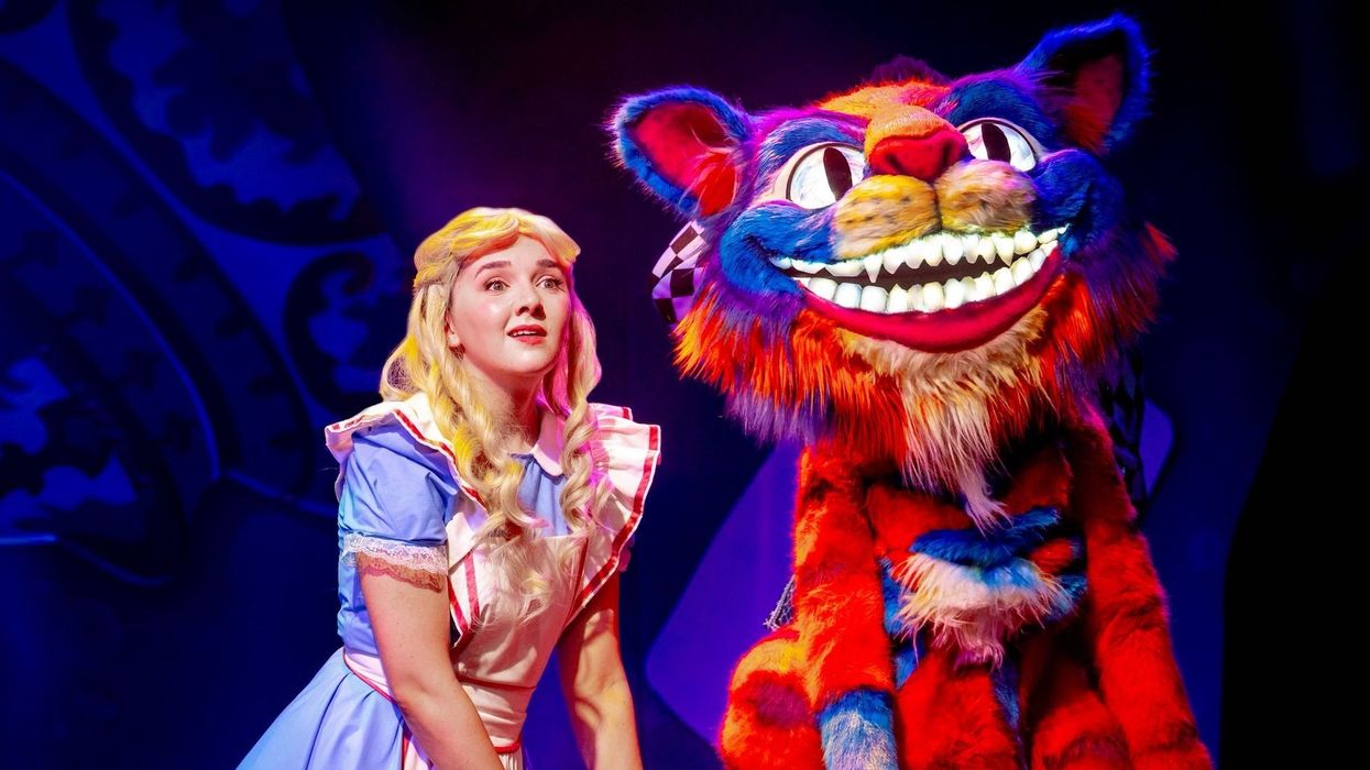 Alice in Wonderland beside a colorful, grinning Cheshire Cat on stage.