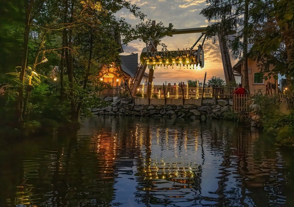 Amusement park ride at sunset over a serene, reflective pond surrounded by trees.