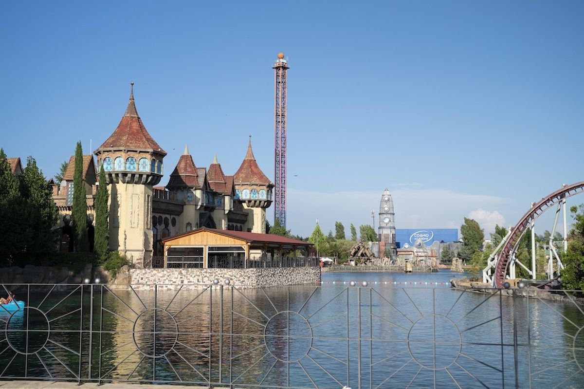 Amusement park with castle-like building, tall tower, and roller coaster by a lake.