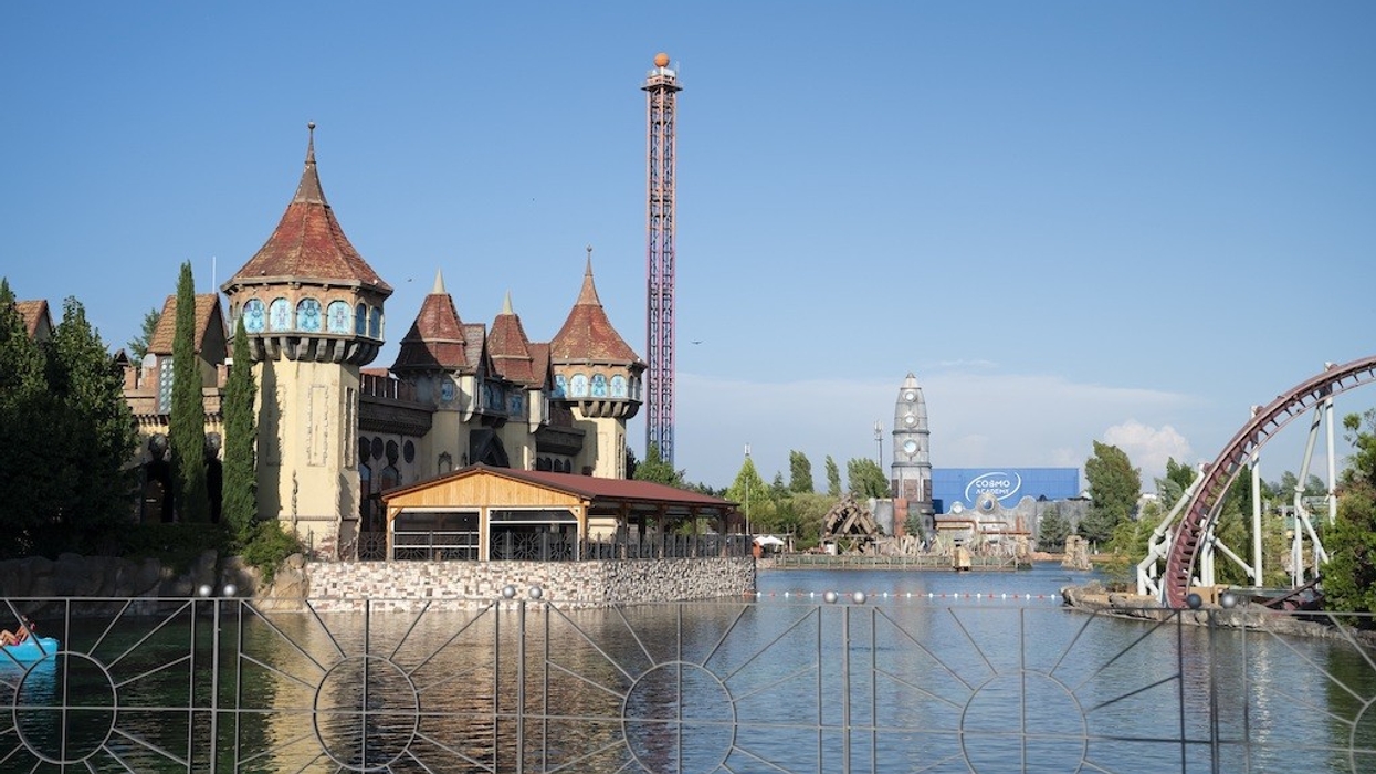 Amusement park with castle-like building, tall tower, and roller coaster by a lake.
