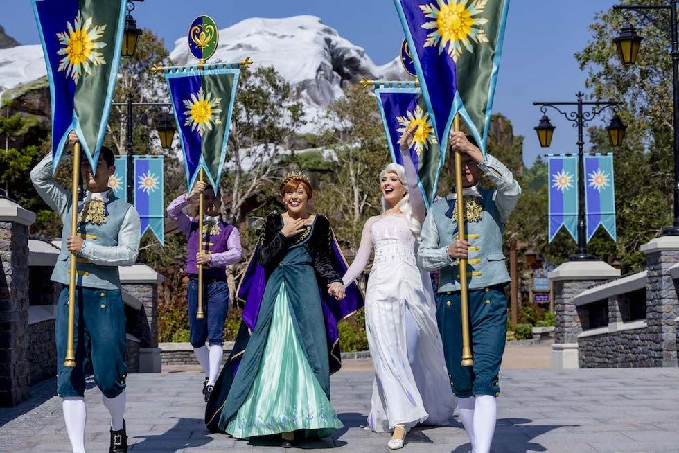 Ana and Elsa characters in festive attire walk with banners in a snowy-themed area, World of Frozen at Hong Kong Disneyland
