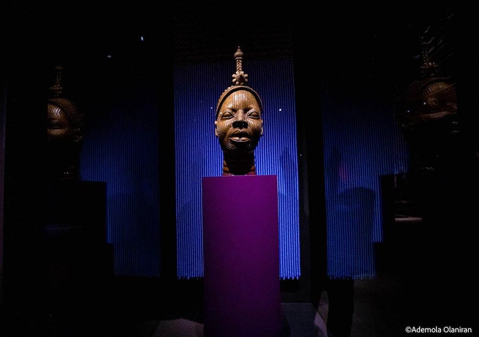 Ancient carved wooden head on purple pedestal with dark, moody lighting.