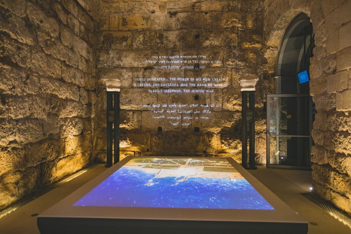 Ancient stone wall with multilingual text and a map display table at Caesarea Harbour Visitor Centre