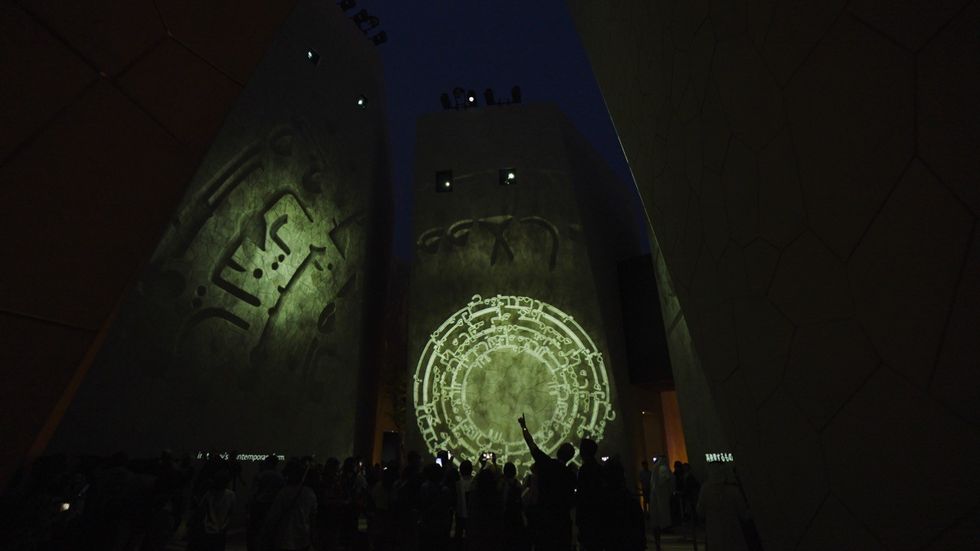 Ancient symbols projected on tall walls, crowd gathered below in a dim setting.