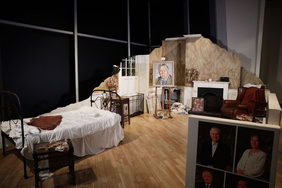 Art studio with bed, paintings, easel, chair, and fireplace in a dimly lit room.