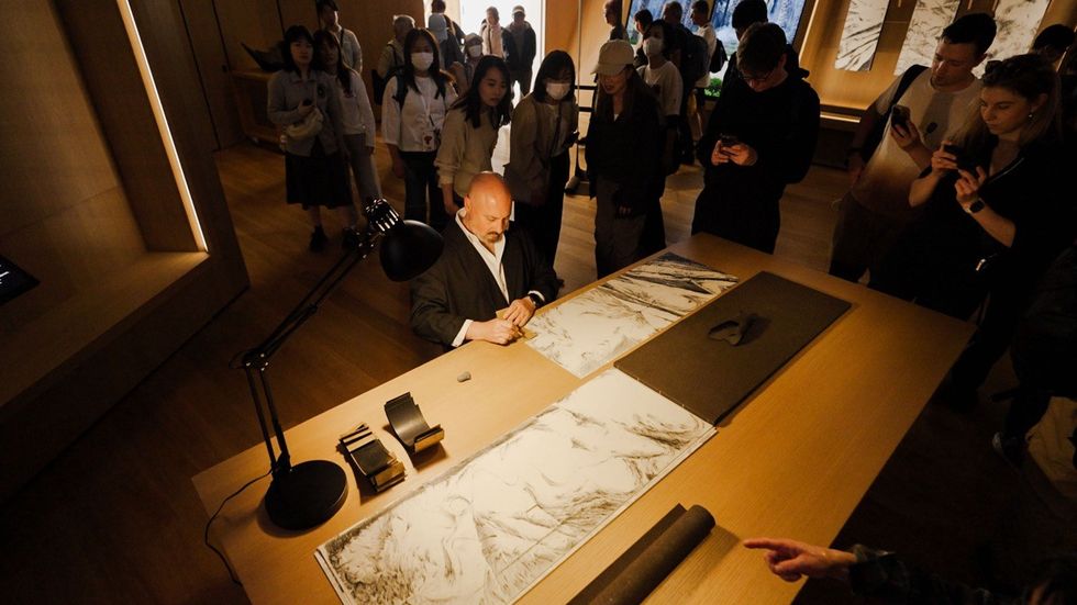 Artist drawing at a table, surrounded by a crowd in a gallery setting.