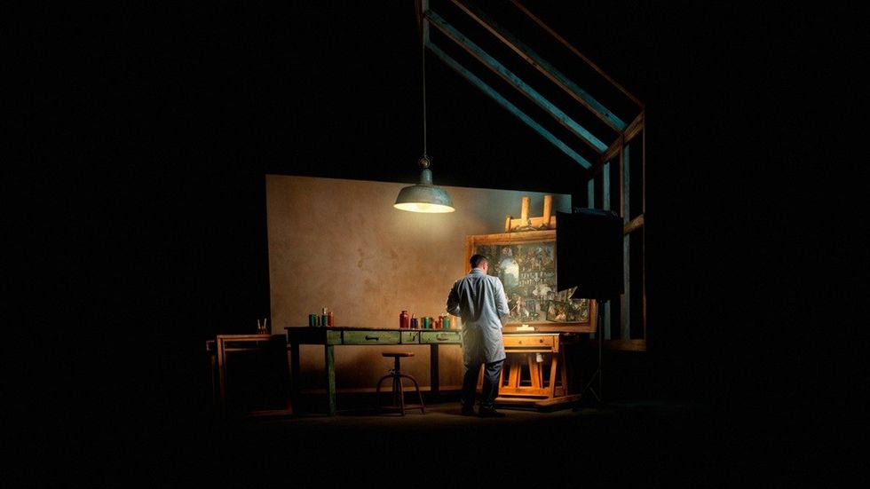 Artist in dim studio with easel and bottles under a hanging lamp.