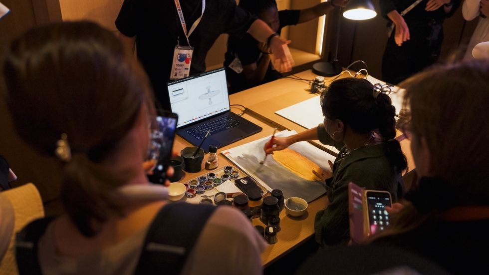 Artist paints at a table, surrounded by onlookers, with laptop and paint pots nearby.