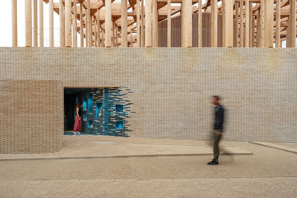 ATELIER BR\u00dcCKNER Uzbekistan Pavilion \u2013 Expo 2025 Osaka Brick wall with blue niches, person walking in foreground, another inside doorway.
