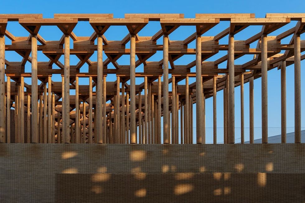 ATELIER BR\u00dcCKNER Uzbekistan Pavilion \u2013 Expo 2025 Osaka Wooden structure with vertical beams casting shadows against a blue sky.