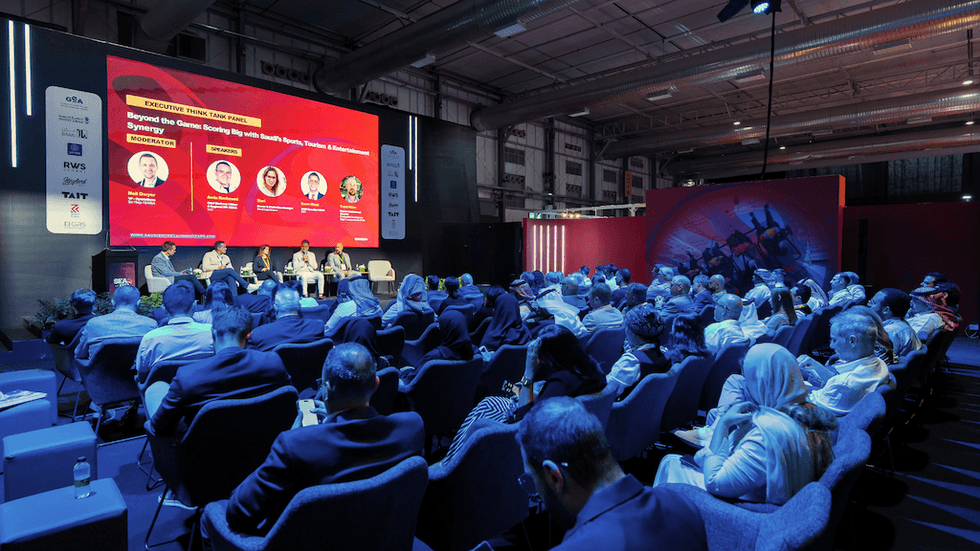 Audience attends a panel discussion at a conference with a large screen display.