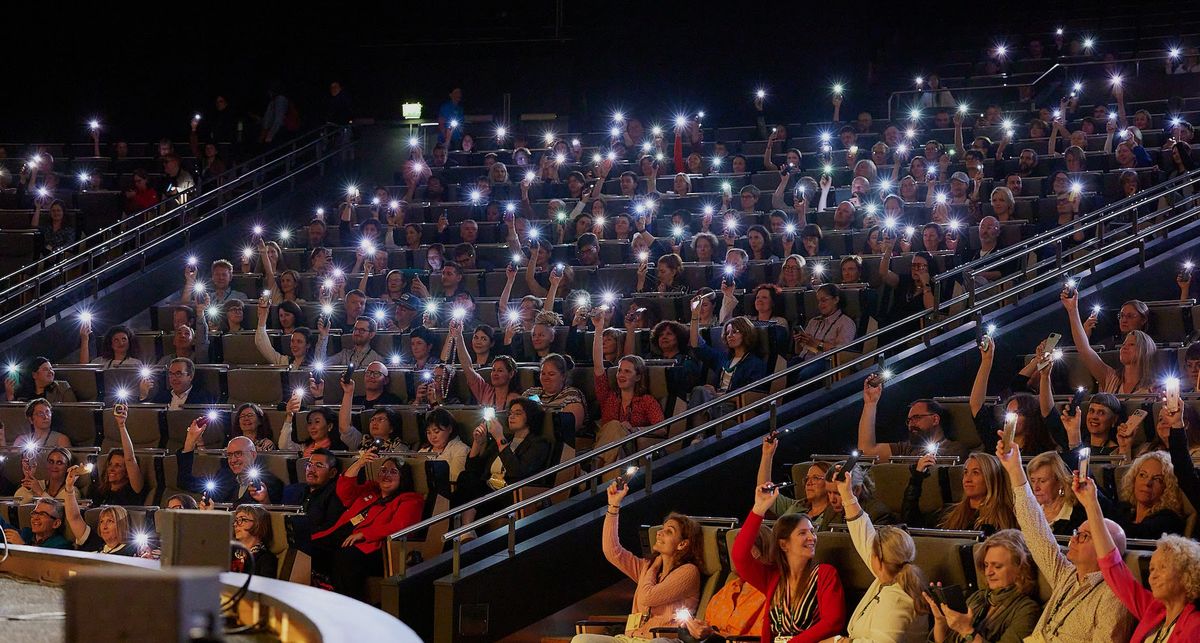 Audience in a theater holding up phones with flashlights on during Ecsite 2026 opening